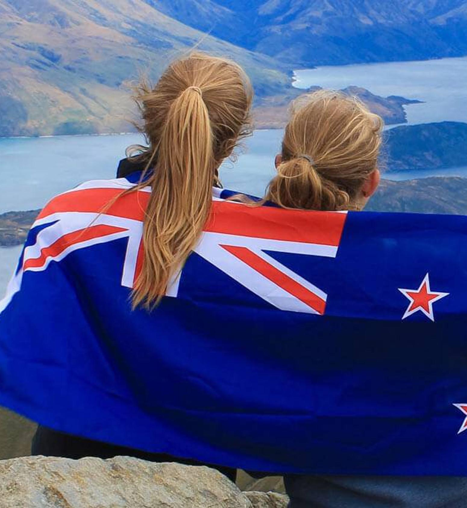 Bild zeigt wie zwei Mädchen in einer Neuseelandflagge eingehüllt, auf einen Berg stehen 