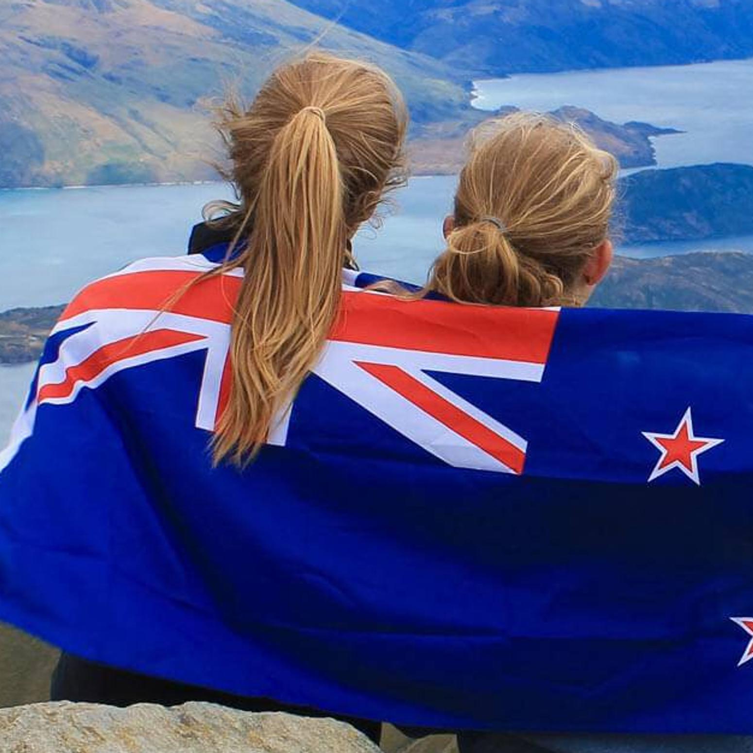 Bild zeigt wie zwei Mädchen in einer Neuseelandflagge eingehüllt, auf einen Berg stehen 