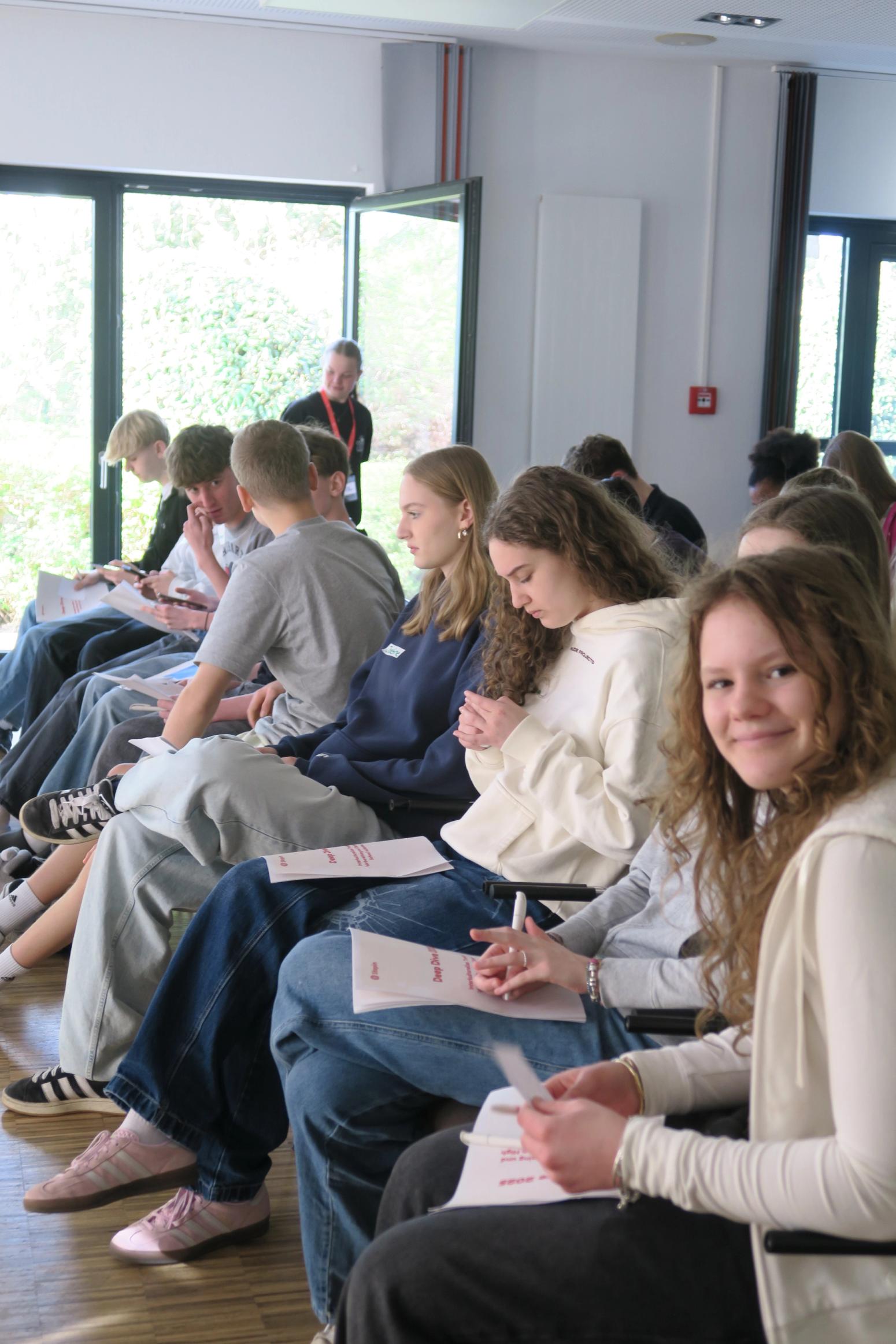 Schülerinnen und Schüler sitzen in einem Seminarraum und schreiben in Hefte; eine Teilnehmerin blickt lächelnd in die Kamera.