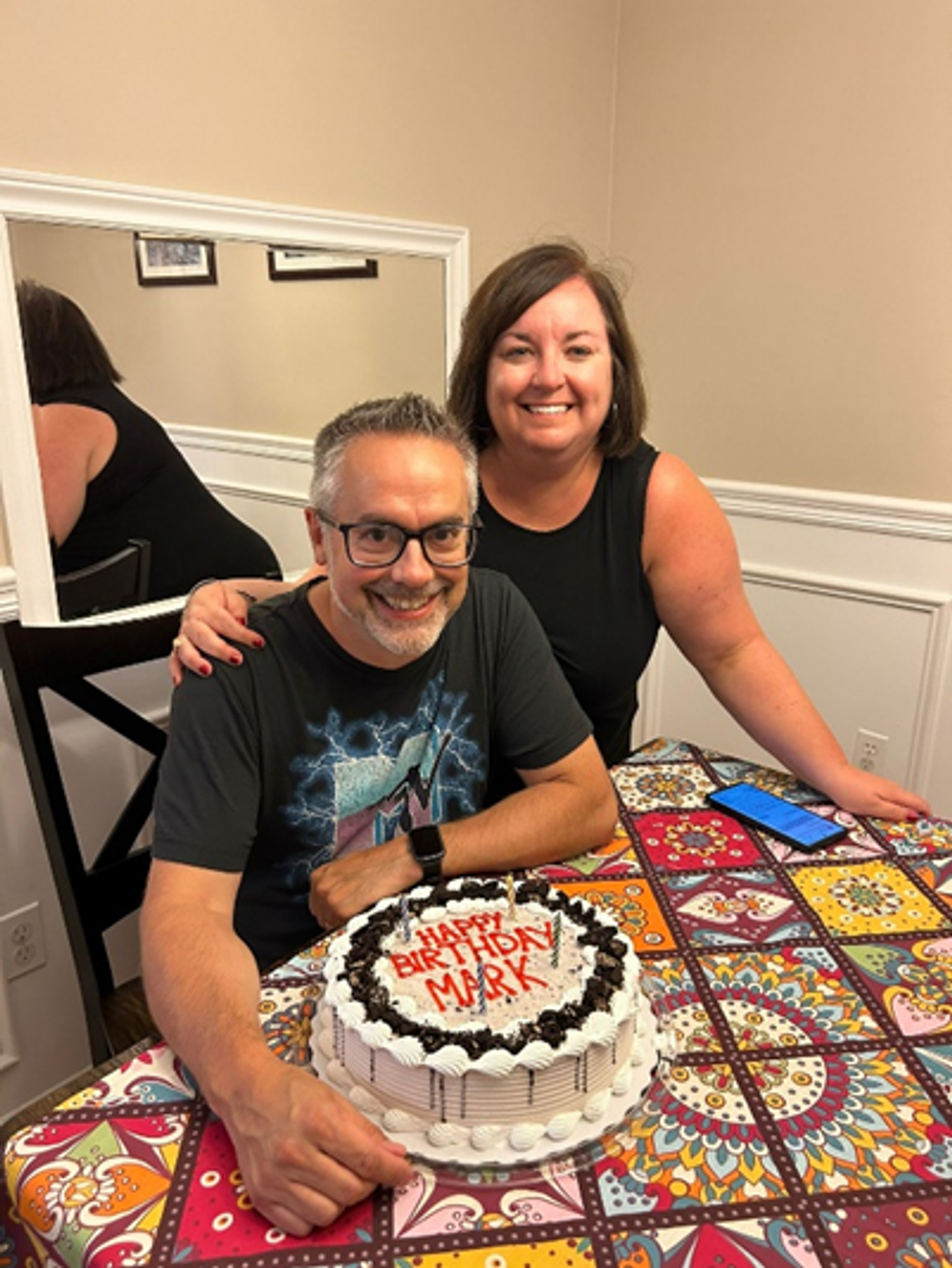 Gastvater sitzt, Gastmutter steht daneben. Vor ihnen ein Tisch mit Geburtstagskuchen und bunter Tischdecke.