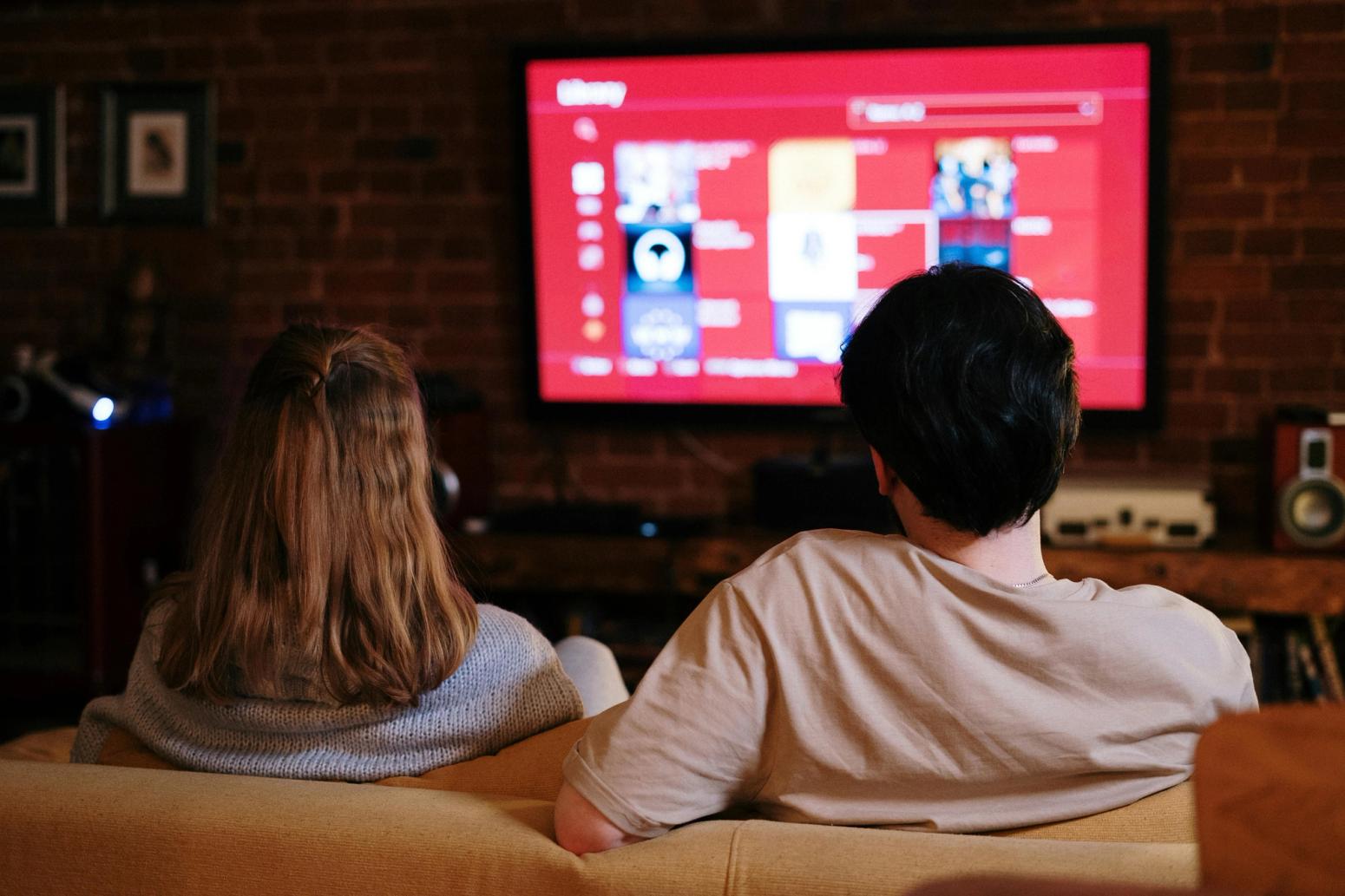 Ein Paar sitzt auf einem Sofa in einem gemütlichen Wohnzimmer mit Backsteinwand und schaut gemeinsam auf einen großen Fernseher, auf dem ein Streaming-Dienst oder eine Medienbibliothek angezeigt wird.