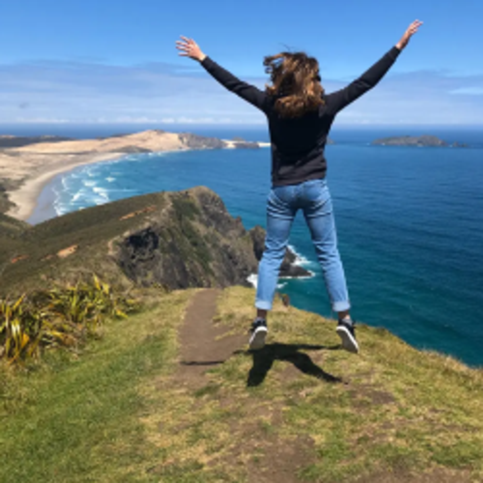 Austauschschülerin springt jubelnd am Küstenpfad in Neuseeland – Blick auf türkisblaues Meer, traumhafte Natur und das Gefühl grenzenloser Freiheit im Auslandsjahr.