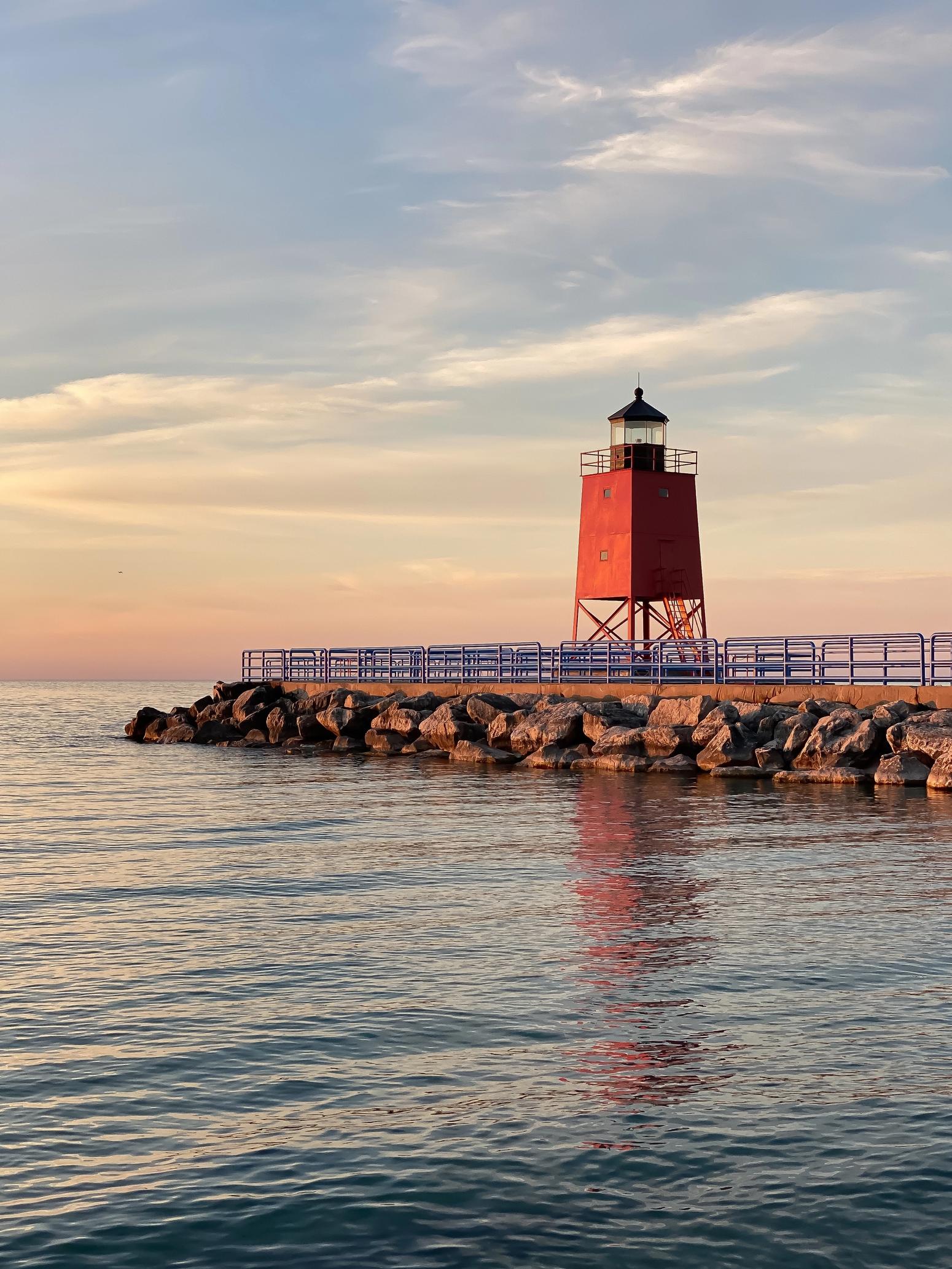 Roter Leuchtturm auf einer Steinmole bei Sonnenuntergang, umgeben von ruhigem Wasser und einem malerischen Himmel mit leichten Wolken – maritimes Panorama am Seeufer.