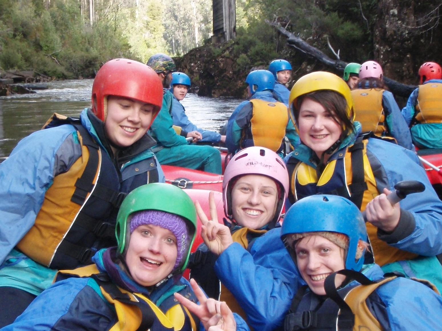 Eine Gruppe junger Menschen trägt Helme und Schwimmwesten, während sie lachend und posierend auf einem Fluss in einem Schlauchboot sitzen. Sie sind von dichtem Wald umgeben, und im Hintergrund ist eine alte Brücke zu sehen.
