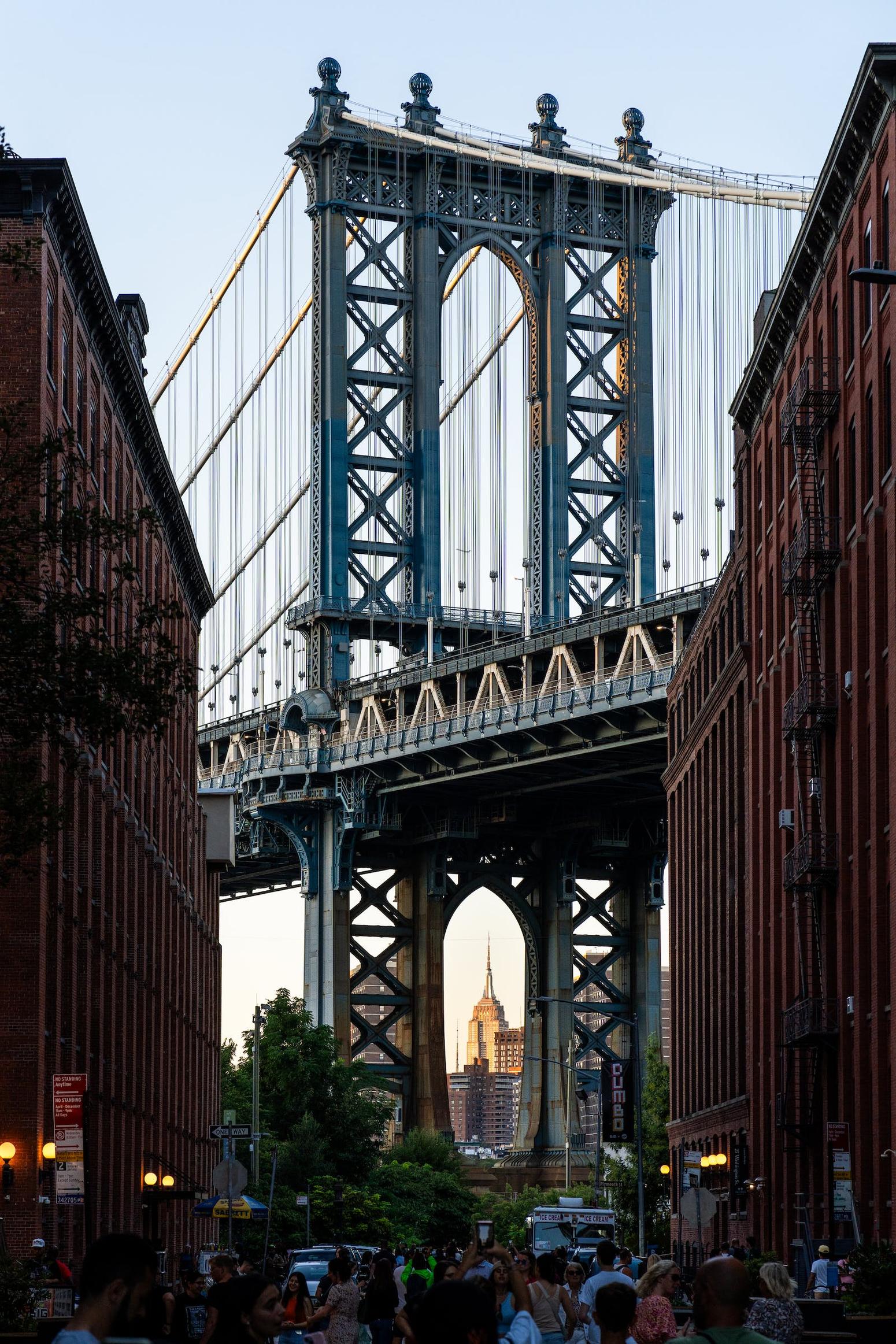 Blick auf die Manhattan Bridge in New York City von einer Straße aus, das Empire State Building ist durch die Brückenbögen zu sehen, umgeben von hohen Backsteingebäuden.