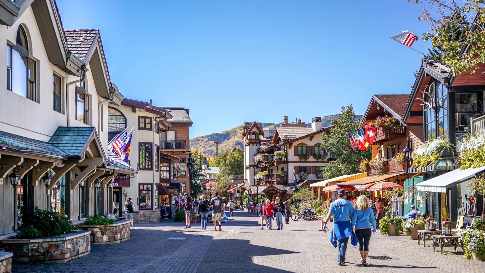 Fußgängerzone in einem alpinen Ferienort mit amerikanischen und internationalen Flaggen, gesäumt von charmanten Gebäuden im Chalet-Stil, bei sonnigem Wetter und klarem Himmel – Touristen schlendern durch das belebte Zentrum