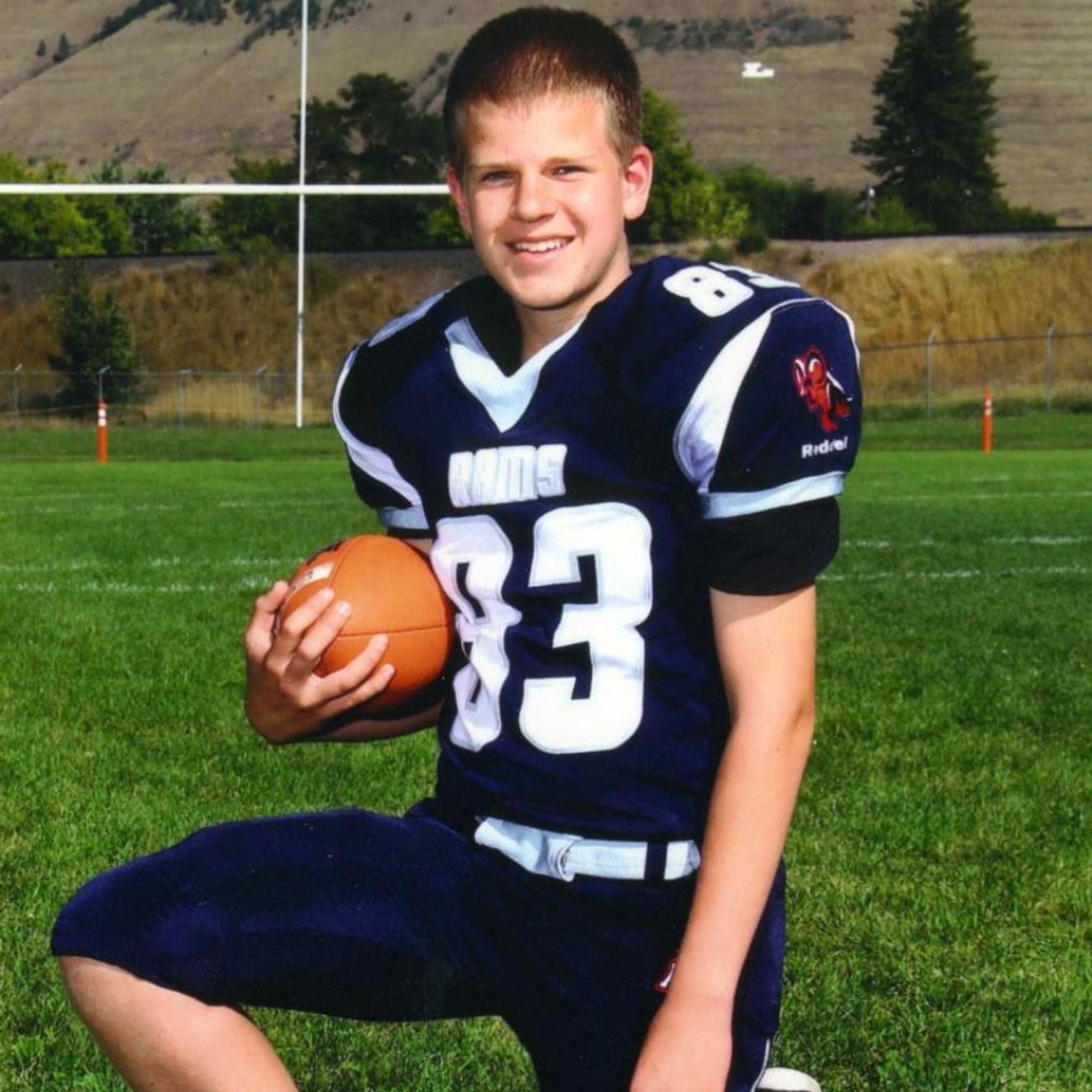 Junge im Footballtrikot kniet auf einem Rasenfeld und hält einen Football, im Hintergrund Torpfosten und hügelige Landschaft.