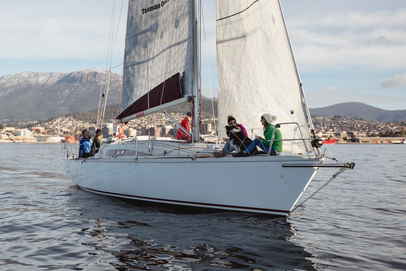 Ein Segelboot gleitet über das ruhige Wasser des Derwent River, an Bord sind mehrere Menschen. Im Hintergrund sieht man die Stadt Hobart und den Mount Wellington unter einem klaren Himmel. Credit: Samuel Shelley.