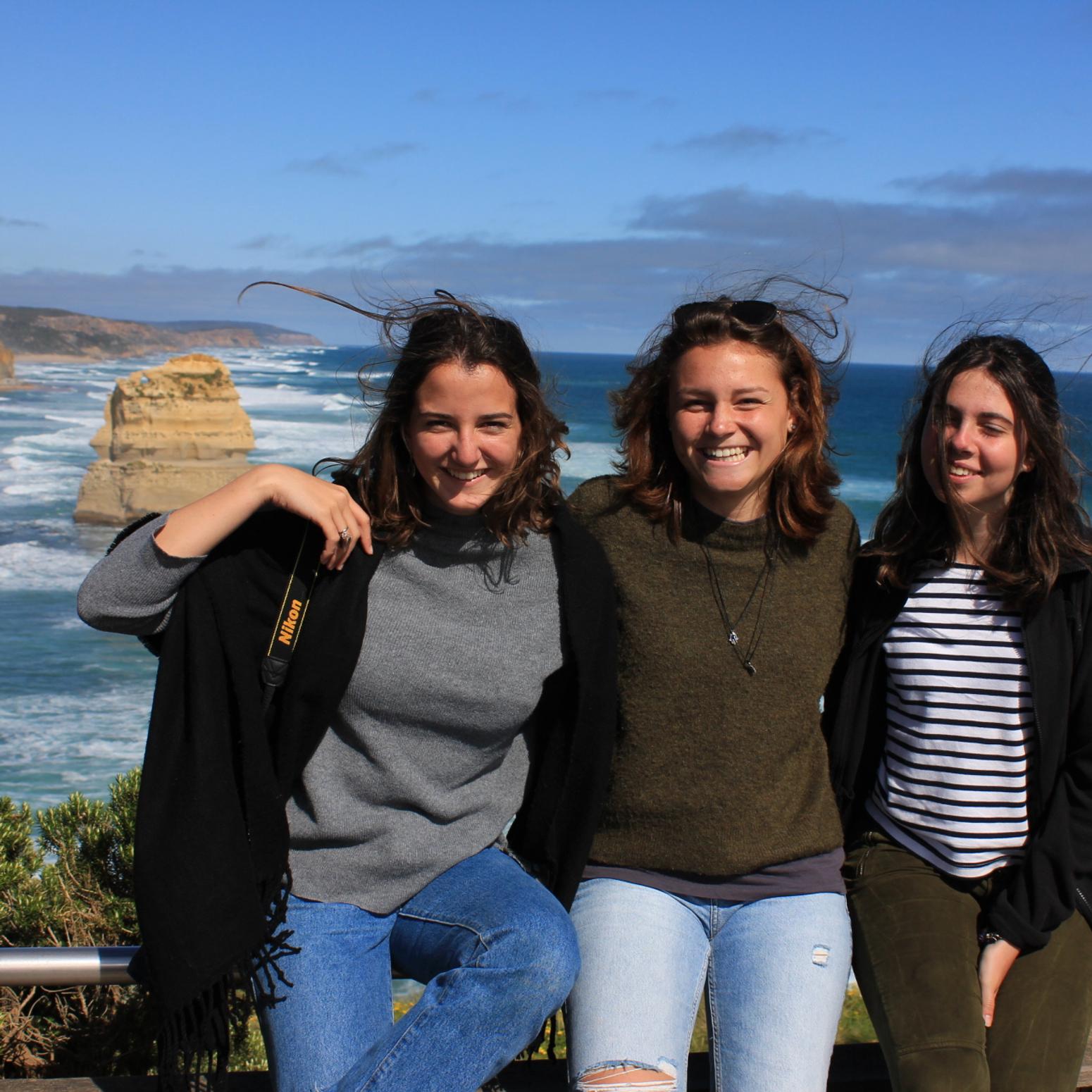 Drei junge Frauen lächeln vor einer Küstenlandschaft mit Meer und Klippen