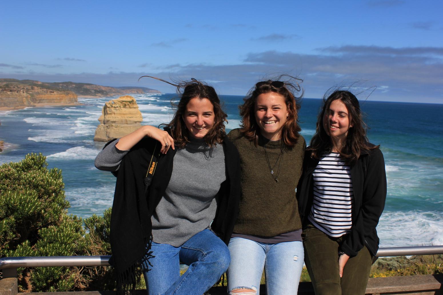 Drei junge Frauen lächeln vor einer Küstenlandschaft mit Meer und Klippen