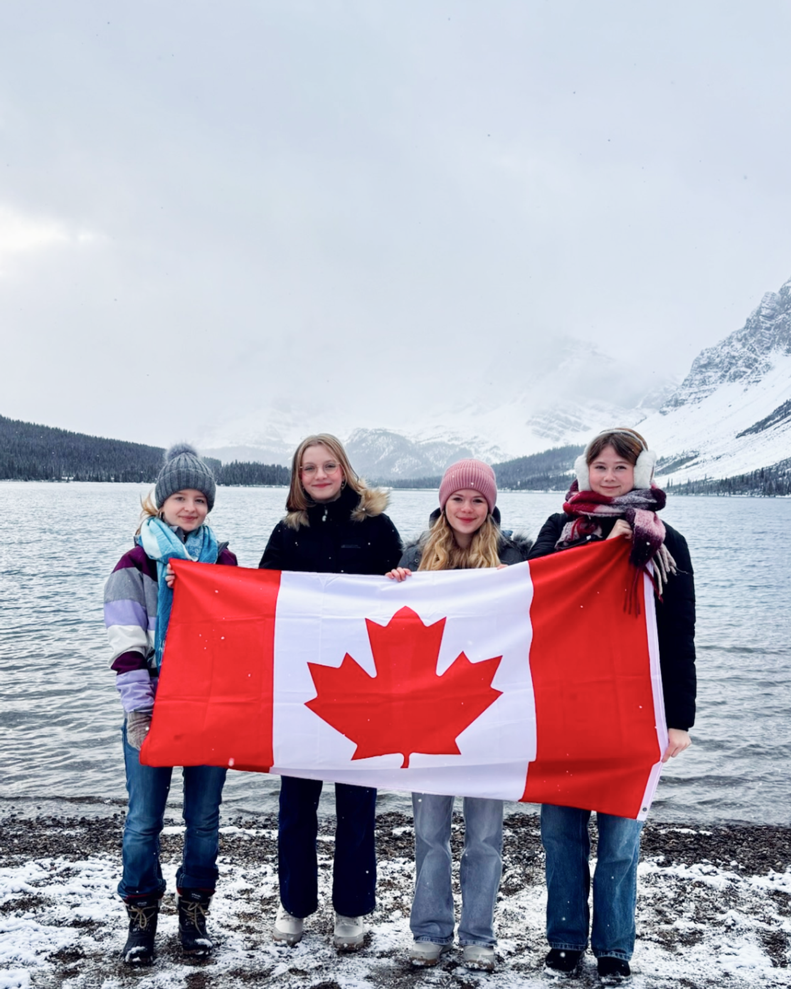 Vier Schüler halten eine Kanada-Flagge vor einem Bergsee im winterlichen Kanada