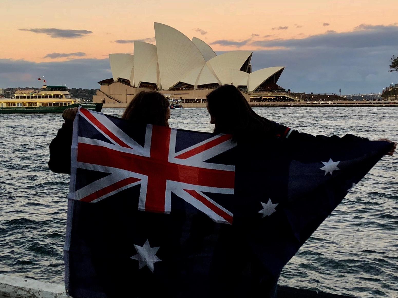 Zwei junge Frauen stehen vor dem Sydney Opera House und halten eine australische Flagge in die Luft