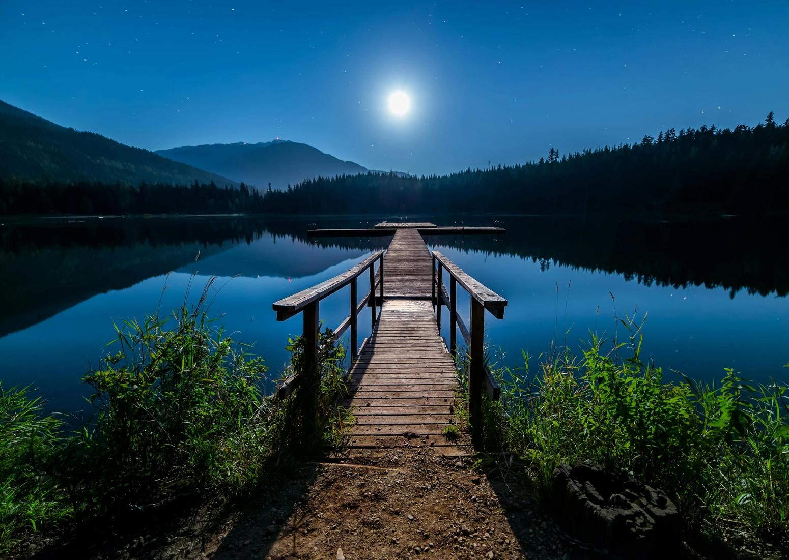 Holzsteg führt auf einen ruhigen, spiegelnden See unter einem leuchtenden Vollmond in einer sternenklaren Nacht.