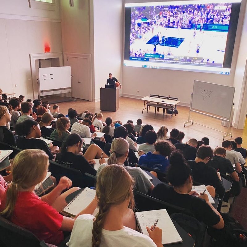 A lecturer addresses a full hall of students watching basketball footage on a large screen.