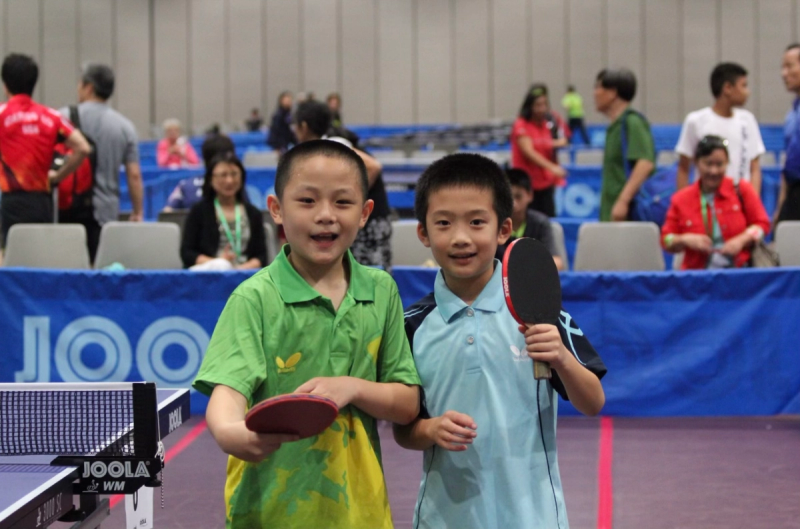 Two young boys in green and blue jerseys hold table tennis bats beside a JOOLA table at a tournament.
