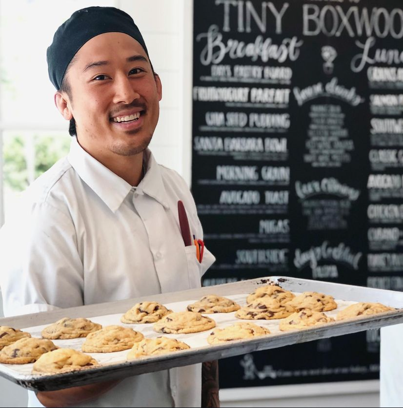 A smiling man in a chef cap holds a tray of fresh cookies in front of a handwritten cafe menu board.