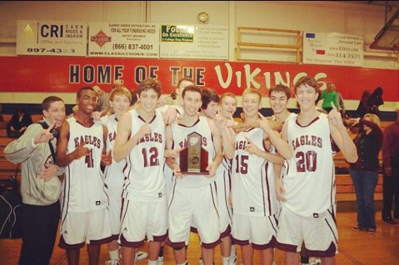 A high school basketball team in Eagles jerseys pose with a trophy under a Home of the Vikings banner.