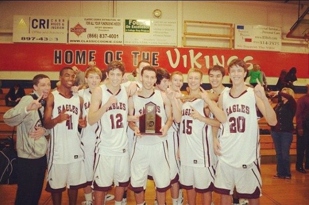 A high school basketball team in Eagles jerseys pose with a trophy under a Home of the Vikings banner.