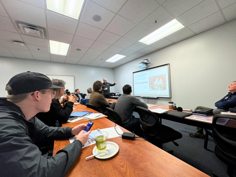 Attendees seated around a conference table watching a presenter beside a projector screen.