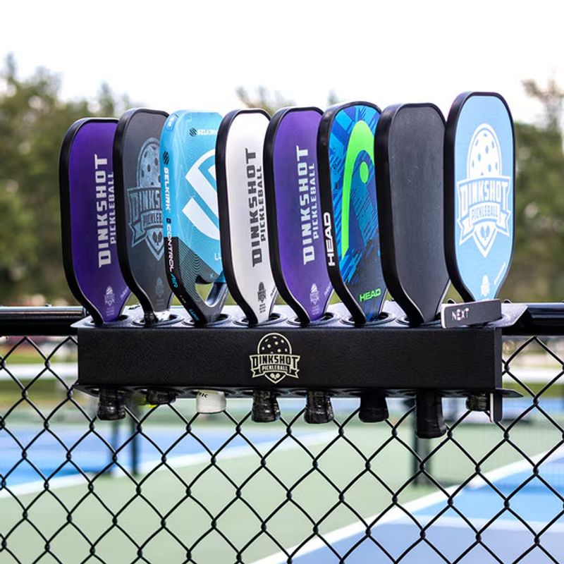Eight paddles from Dinkshot, Head and others racked along a chain-link fence beside a court.