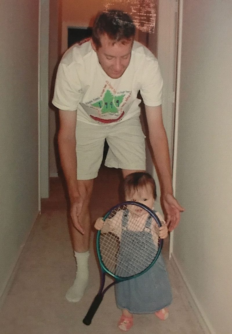 Old photograph of a man steadying a toddler who is holding a full-size tennis racquet in a hallway.