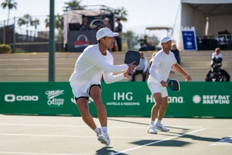 Two players in white lunge for a shot on a green outdoor court lined with sponsor banners.