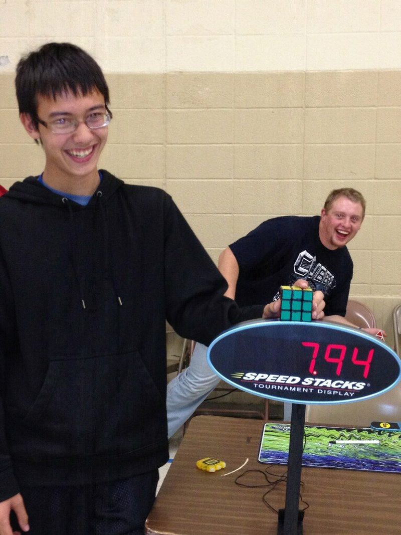 A smiling teenager beside a Speed Stacks tournament display reading 7.94 seconds as a friend holds a Rubik cube.