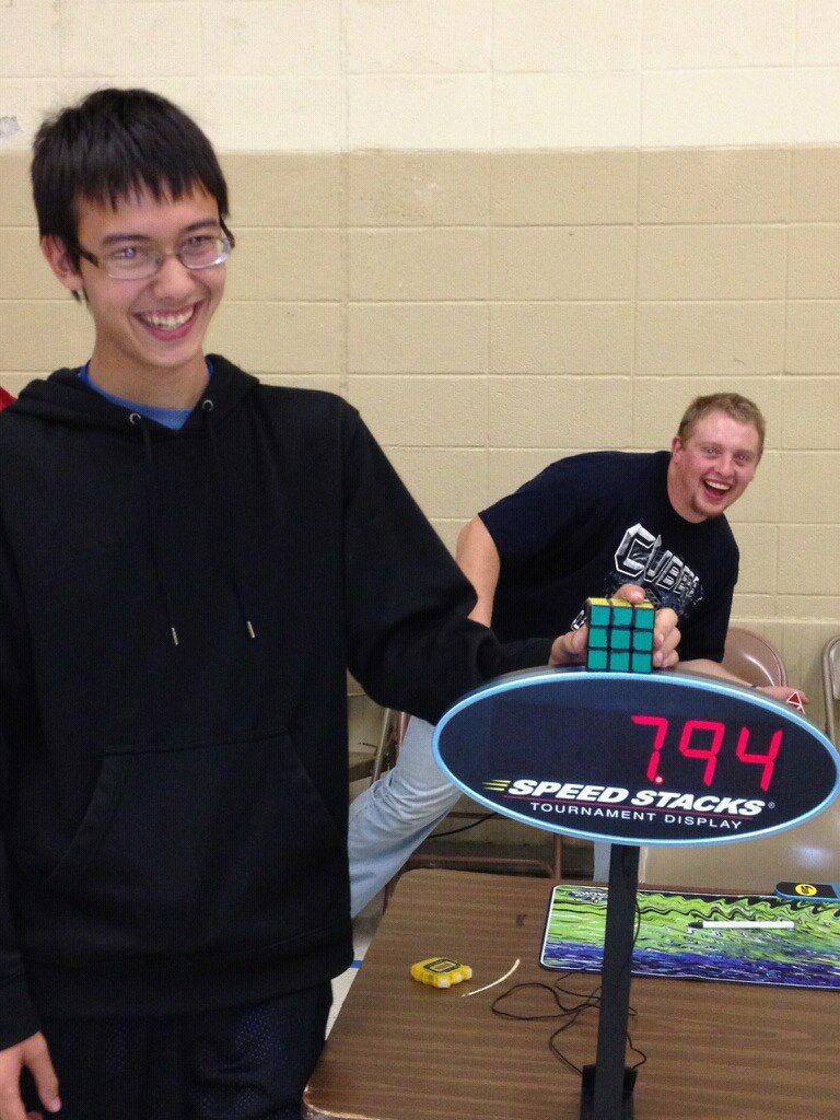 A smiling teenager beside a Speed Stacks tournament display reading 7.94 seconds as a friend holds a Rubik cube.