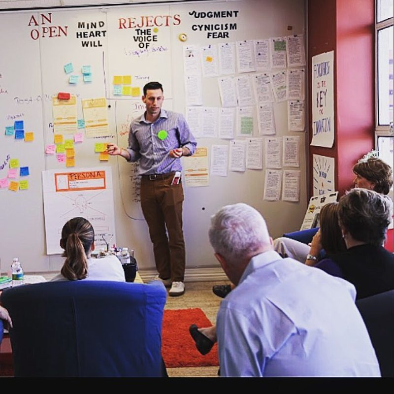 A man presents to a seated group in front of a whiteboard covered in sticky notes and printed sheets.