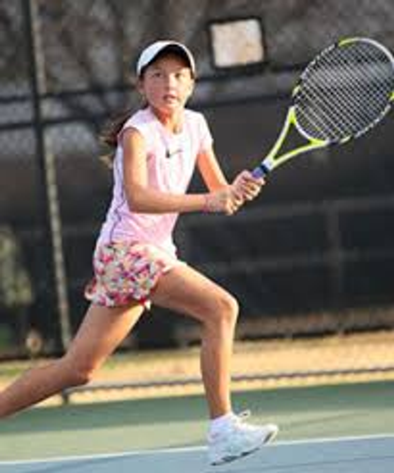 A girl in a pink top and white visor follows through on a two-handed tennis backhand on an outdoor court.