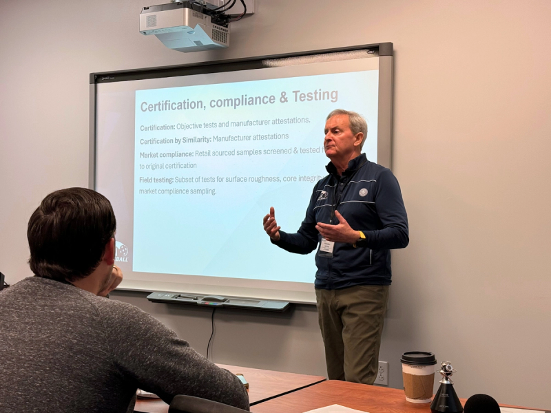 A presenter in a USA Pickleball jacket speaking beside a slide headed certification, compliance and testing.