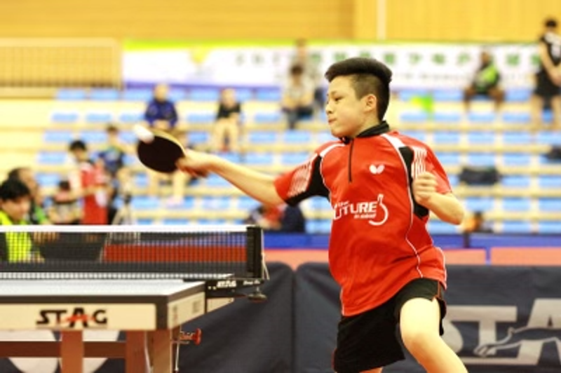 A young player in a red jersey plays a forehand at a table tennis table.