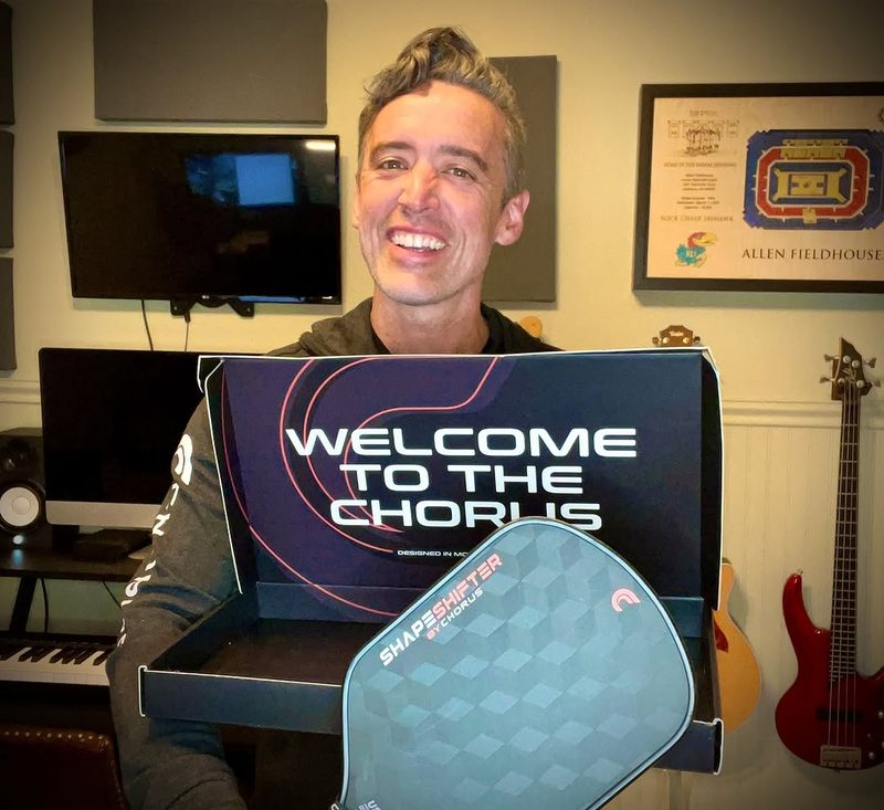 A smiling man holds a Chorus box reading welcome to the chorus, a Head Shapeshifter paddle propped in front of it.