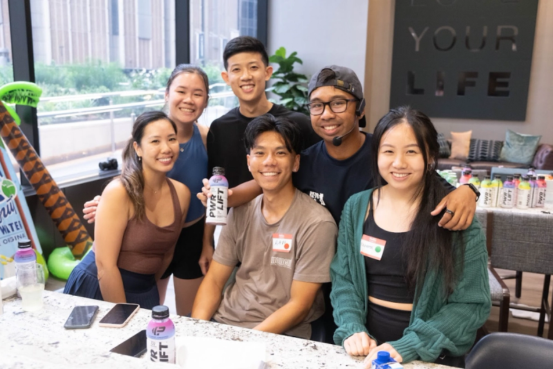 Six friends smiling around a table at an office event, holding drinks, beneath a wall sign reading your life.