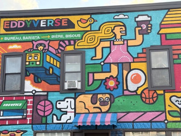 A colorful street mural titled "Eddyverse" by artists Chris Soueidan (Loogart) and Phil Landry on Rue Eddy in Gatineau. Designed in Loogart’s signature playful illustration style, the mural depicts a vibrant mini-universe filled with coffee-shop-inspired elements, cartoon characters, and neighborhood pop culture references painted across an exterior wall.