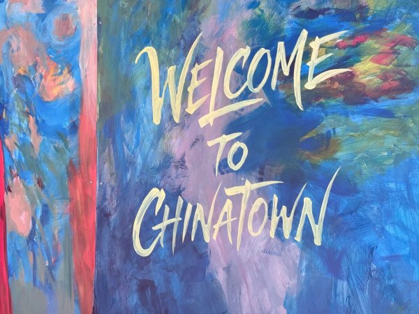 A vibrant outdoor mural painted on a long wall next to a wooden deck, featuring a stylized figure in a traditional Chinese opera headdress and the words "Welcome to Chinatown" written in gold cursive on a blue background.