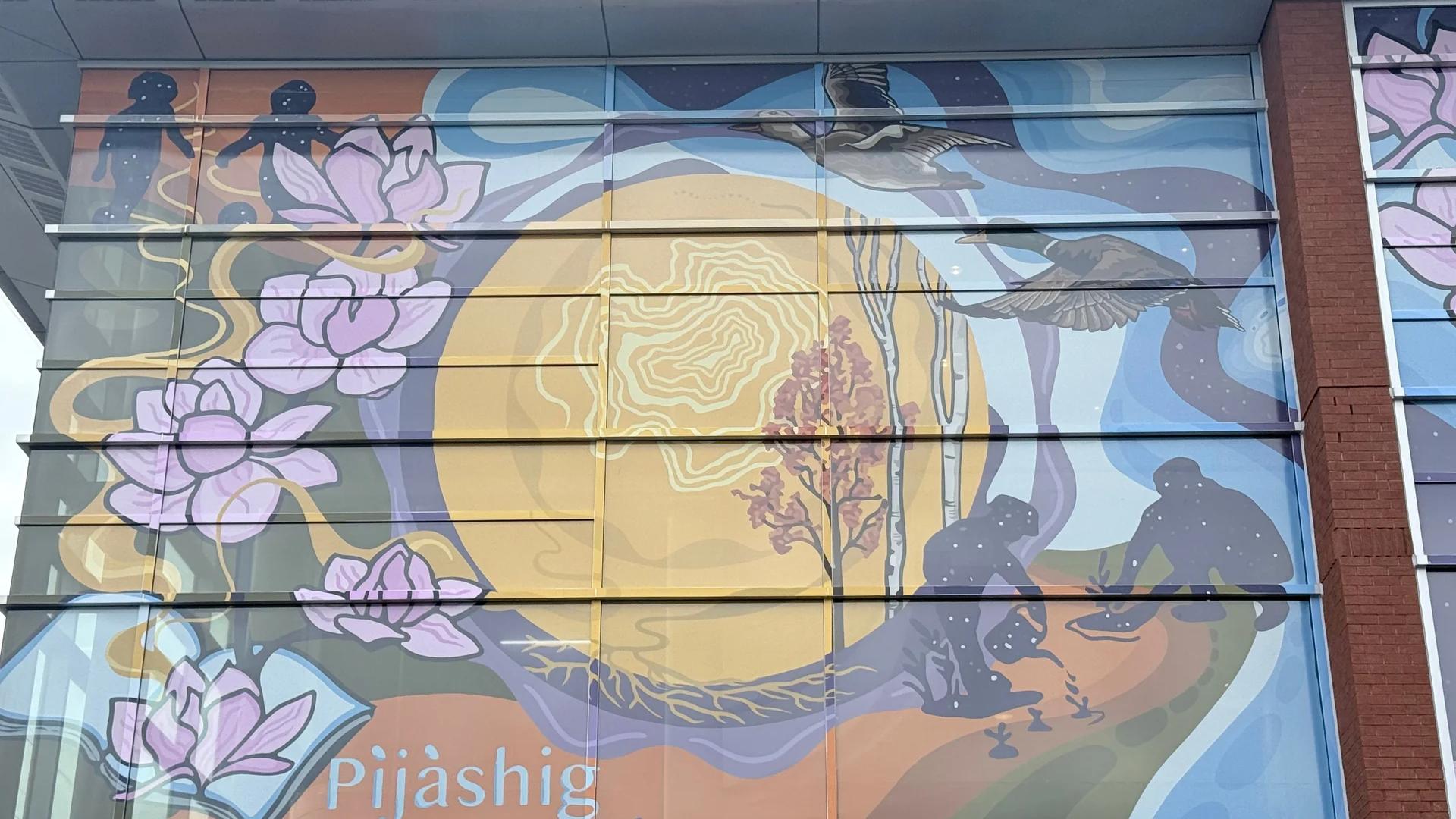 Colorful mural on a building displays pink lotus flowers, flying birds, star-filled figures, and 'Pijàshig Welcome' on Chamberlain Avenue, Ottawa.