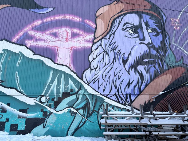 Large-scale outdoor mural by Ottawa artists Dems and Doll in Centretown, depicting a bearded figure reminiscent of Leonardo da Vinci alongside the Vitruvian Man and dynamic water imagery, painted on a building exterior in winter