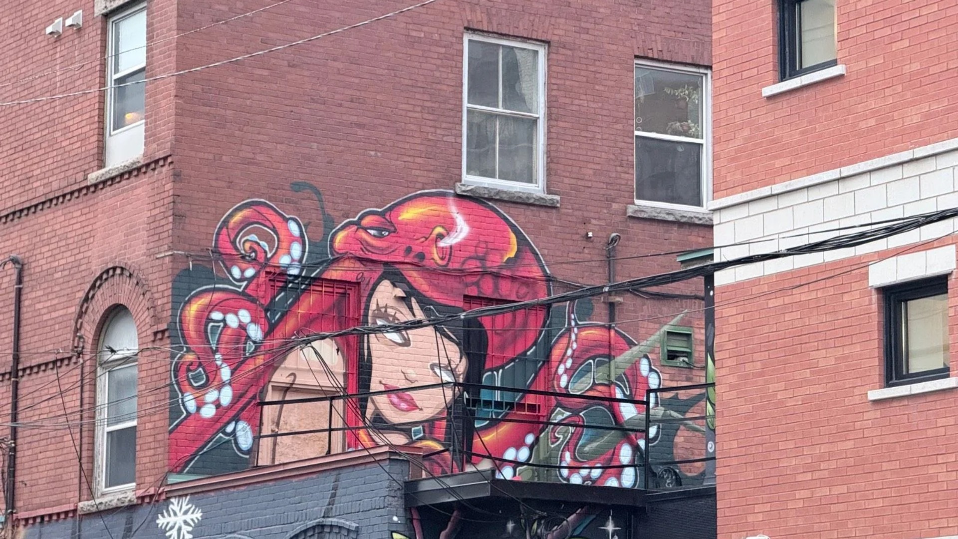 Vibrant mural of a woman with red octopus tentacles on a brick building, traversed by utility wires, Lewis Street, Centretown, Ottawa.