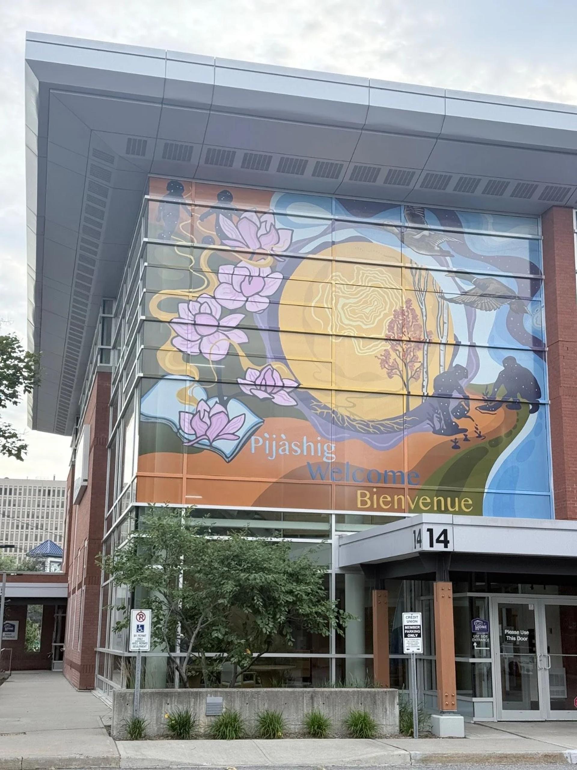 A vibrant mural on a glass building depicts lotus flowers, a sun, planting figures, and 'Pijàshig Welcome Bienvenue' on Chamberlain Avenue, Capital, Ottawa.