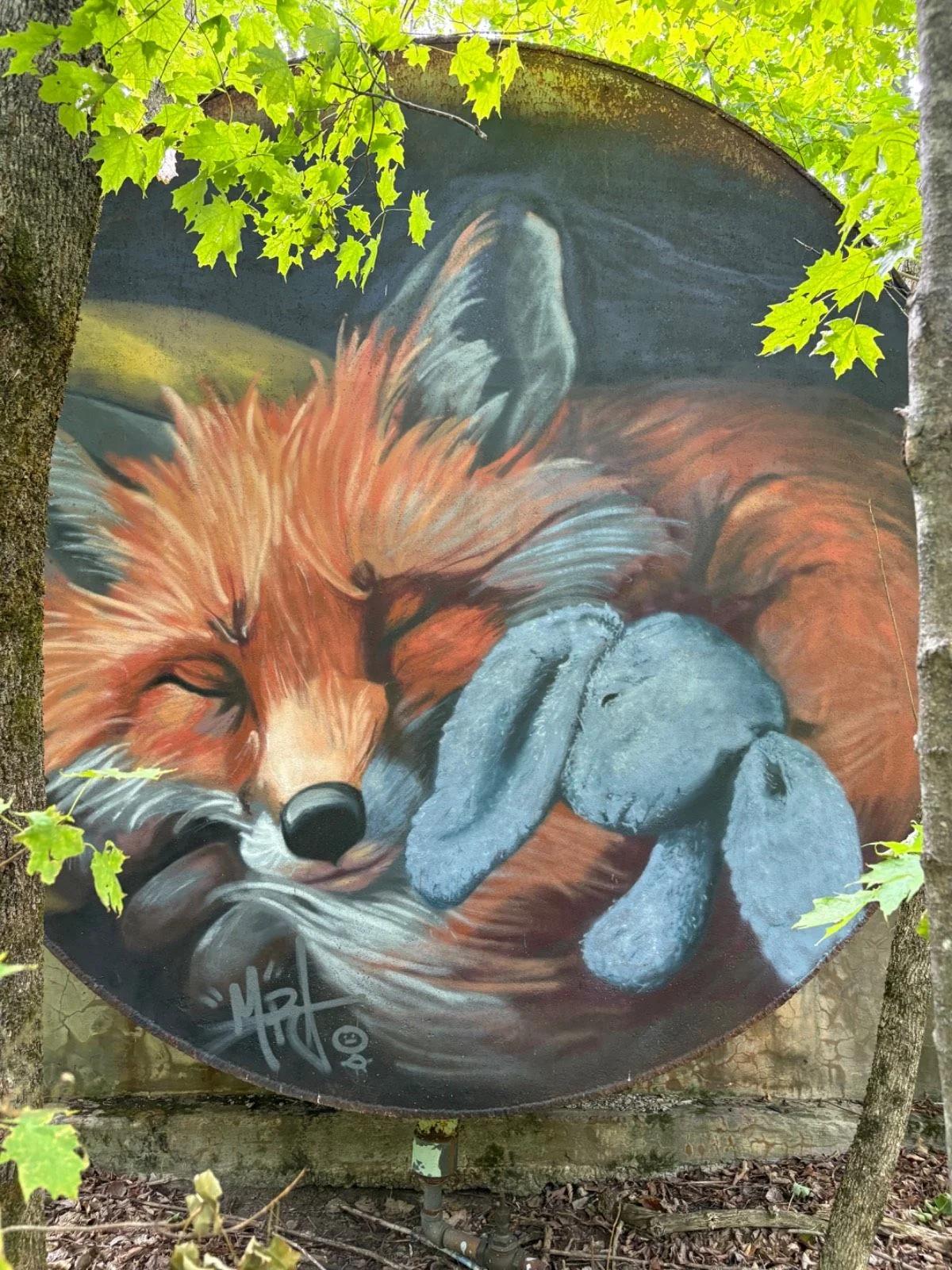 Vibrant mural on round rusty metal shows a sleeping orange fox with a blue bunny, framed by trees and leaves, Trail #24, Pontiac, Ottawa.