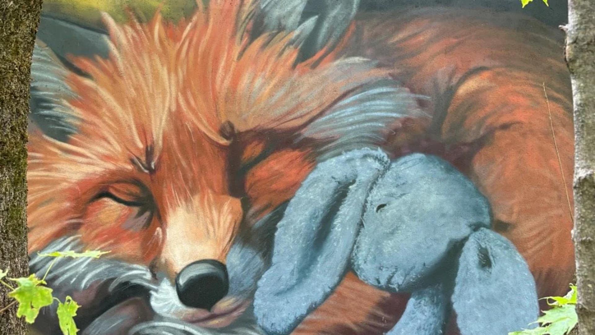 Vibrant mural on round rusty metal shows a sleeping orange fox with a blue bunny, framed by trees and leaves, Trail #24, Pontiac, Ottawa.