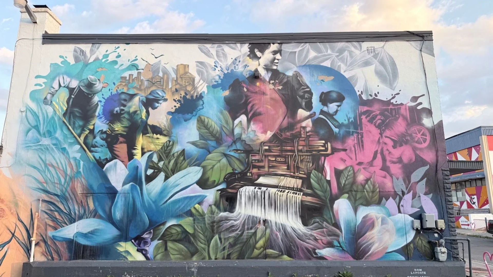 Vibrant mural showing laborers, a city, a loom with thread waterfall, and blue flowers, 148 Rue Eddy, Hull, Gatineau, Ottawa.