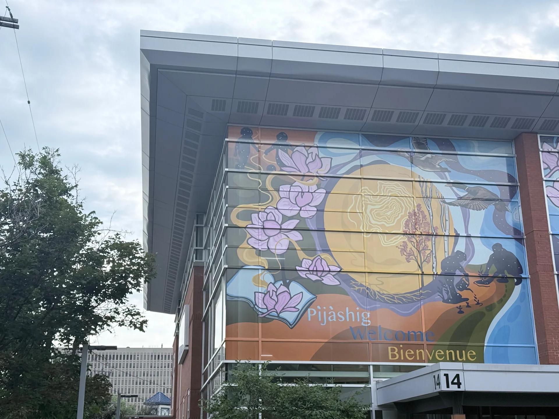 A vibrant mural on building windows depicts stylized lotus flowers, figures, and geese alongside "Pijàshig Welcome Bienvenue" text, Chamberlain Avenue, Capital, Ottawa.