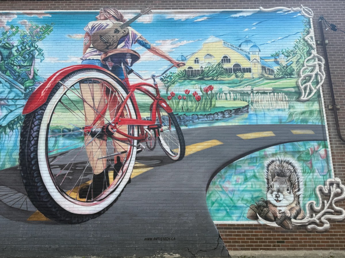 Groovy Glebe mural by Jocelyn Galipeau, showing a person walking a red vintage bicycle with a guitar strapped to their back down a path toward the Aberdeen Pavilion at Lansdowne Park, with red tulips and a squirrel painted in the corner, on a brick wall on Bank Street in the Glebe, Ottawa.