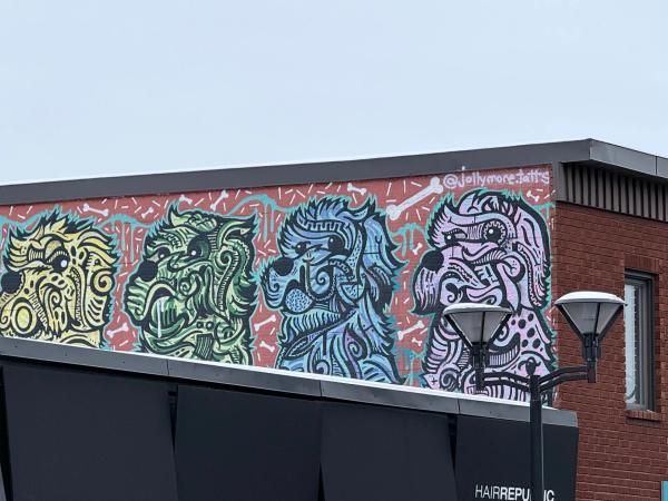 Stylized street art mural of green and black dog heads by Ottawa artist Lester Jollymore on a red brick textured background.
