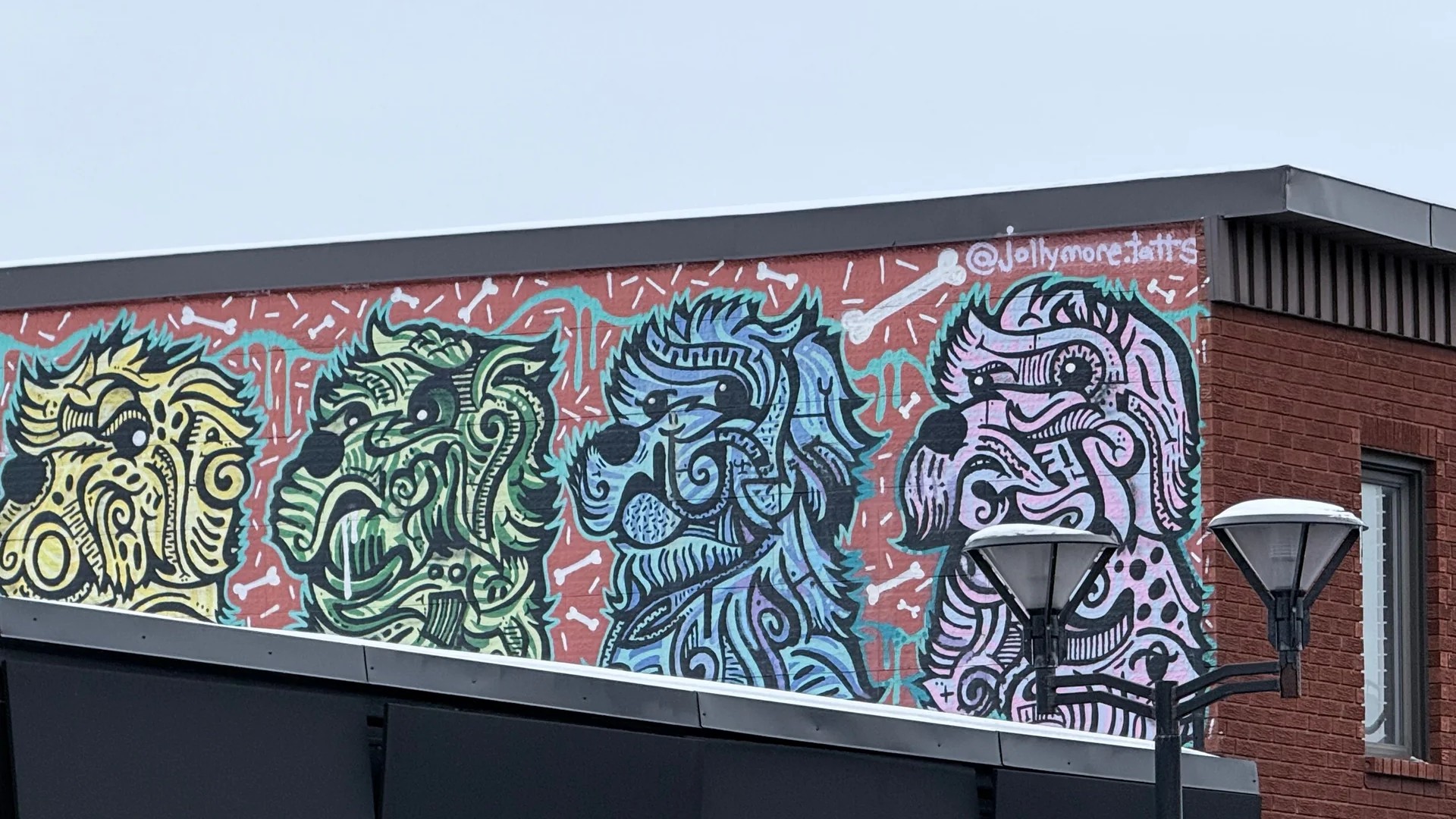 Stylized street art mural of green and black dog heads by Ottawa artist Lester Jollymore on a red brick textured background.