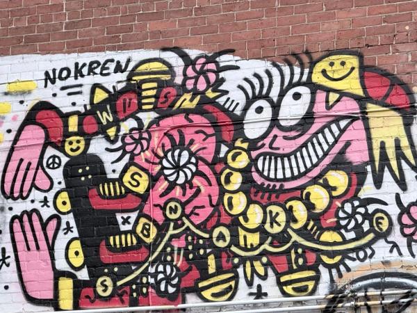 A colourful, cartoon-style mural by Nokren with a grinning pink character, hands, and letters on a brick wall, Centretown, MacLaren Street, Ottawa.