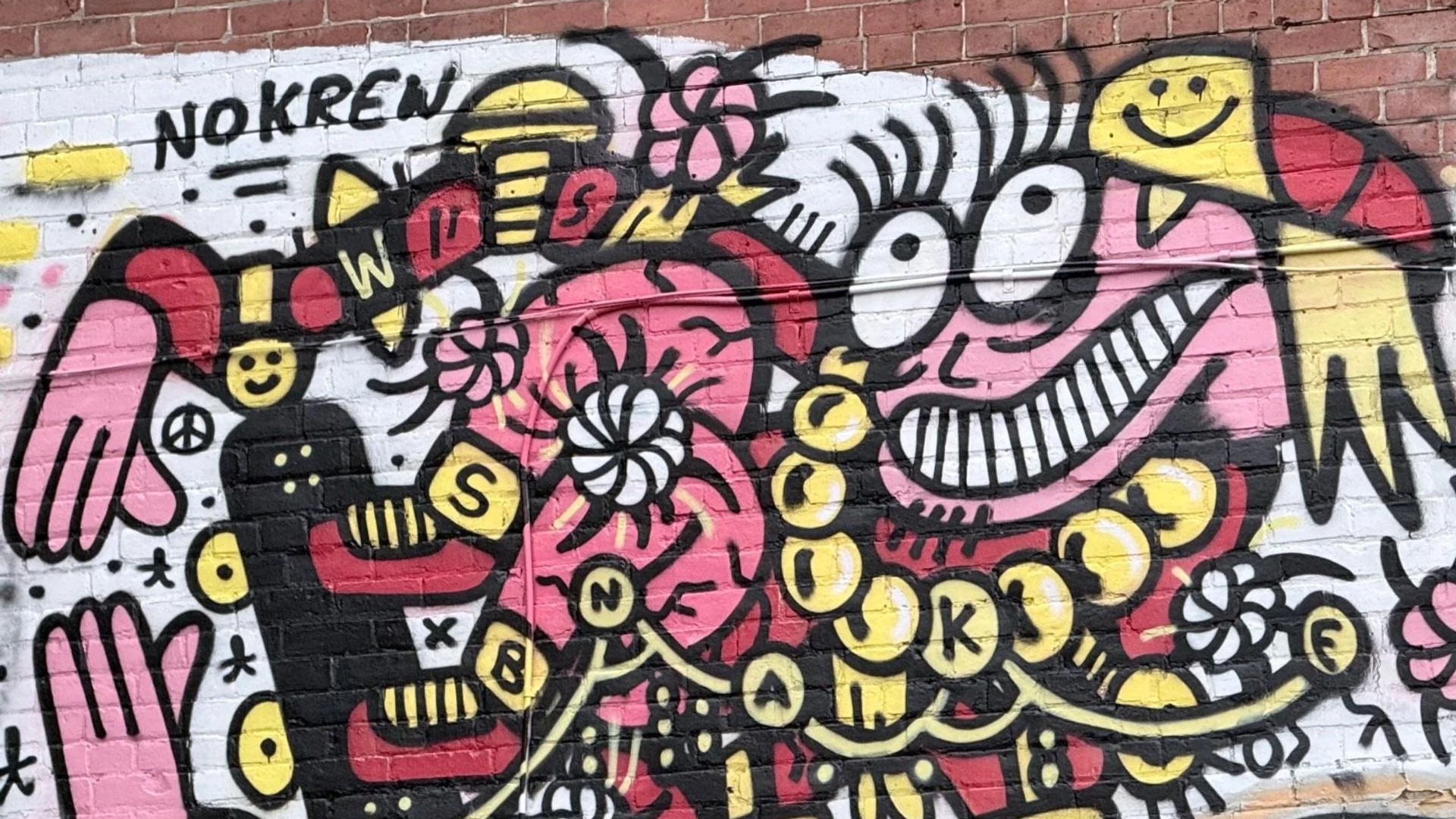 A colourful, cartoon-style mural by Nokren with a grinning pink character, hands, and letters on a brick wall, Centretown, MacLaren Street, Ottawa.