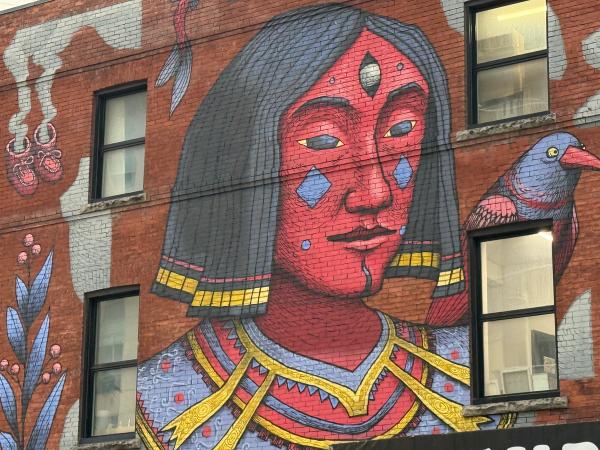 SbuOne mural in Lowertown Ottawa featuring a red-faced, blue-eyed figure with a third eye and dark hair, wearing stylized armor, and a blue and red bird perched on its shoulder, painted on a red brick wall.